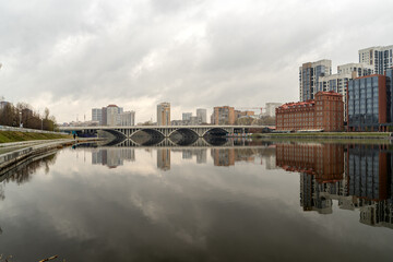 Obraz premium Panoramic view of the river in central Yekaterinburg with Bazhovsky Bridge in the distance. Cloudy sky and autumn or spring season create a moody atmosphere.
