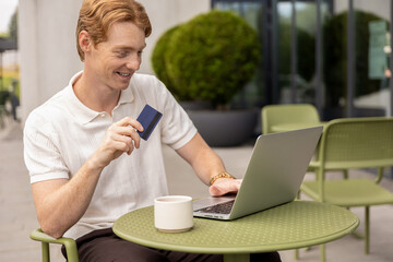 Ginger male freelancer holding credit card typing on laptop paying