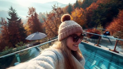 A woman in a beige beanie and sunglasses takes a selfie on a glass observation deck overlooking a scenic landscape.