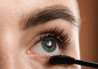 Young woman applying mascara onto her eyelashes on brown background, closeup