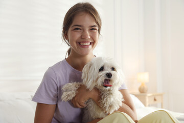 Happy teenage girl with cute Maltese dog on bed at home