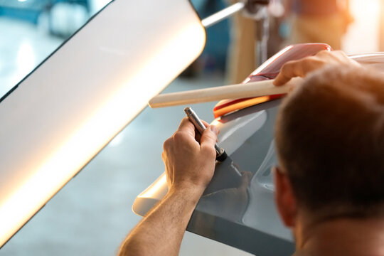 Inside a Garage, an automotive technician is shown from the shoulders up using a specialized tool to repair a dent on a gray automobile. A wooden brace and light are used