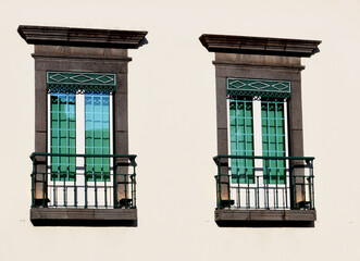 Two traditional windows with green shutters and small balconies on a white wall, architectural detail.