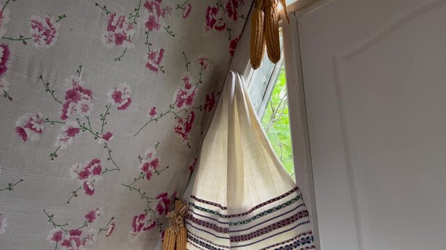 Traditional Ukrainian rushnyk hanging by an open window in a rustic home. Floral wallpaper and hanging corn cobs are visible. A lush summer view outside the window.