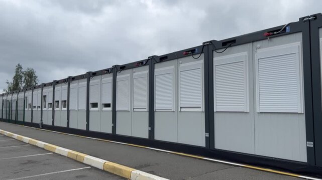  Long modular building with closed roller shutters. Modern container block structure lined up in a row on a parking lot under cloudy skies.