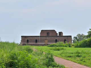 A single-story brick building set in a grassy area, with a narrow brick path leading to it. The building has a rectangular shape and features a central doorway with a wooden door, flanked by evenly sp