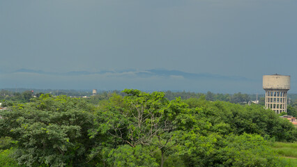 Dense green foliage dominates the foreground, with a robust concrete water tower on the right side. The sky is overcast, contrasting with the lush greenery below. In the distance, a mountain range add