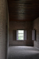 A dimly lit room with brick walls and a wooden ceiling. Two windows with white shutters allow light to enter.