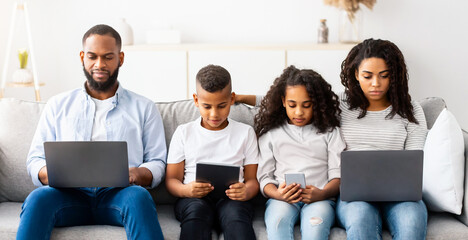 Gadgets Addiction. Portrait of African American family of four people holding and using different electronic devices while sitting on the sofa in living room at home, parents and their little children