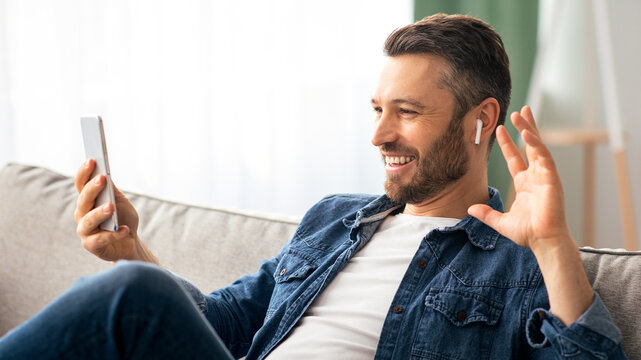 Cheerful bearded man waving at mobile phone while resting on couch at home, side view, copy space. Happy middle-aged man having video conference with friends or relatives, using smartphone and earbuds