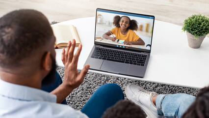 Over the shoulder view of young black family using pc laptop computer with blank empty screen for mock up template, sitting on the sofa in living room at home, waving to webcam, having videocall