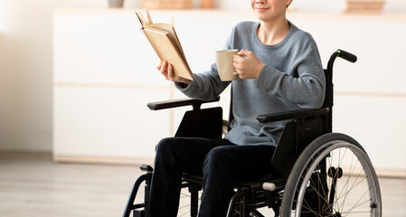 Fototapeta premium Positive impaired teenager in wheelchair drinking tea and reading interesting book at home. Happy handicapped adolescent relaxing, enjoying hot beverage and cool story indoors