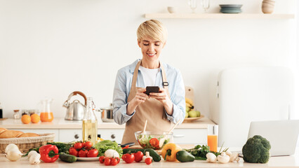 New recipe for healthy dish, health care and hobby, home alone. Happy adult woman blogger in apron prepared salad and typing on smartphone in kitchen interior with bright vegetables on table and juice