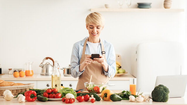 New recipe for healthy dish, health care and hobby, home alone. Happy adult woman blogger in apron prepared salad and typing on smartphone in kitchen interior with bright vegetables on table and juice - Powered by Adobe