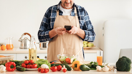 Online chat, dinner invitation, recipe app and technology at home during covid. Smiling senior man in apron writes message while cooking salad in kitchen with table with many fresh vegetables