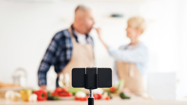 Blogger tasting food, family teaching people to cook healthy eat and shoot video on smartphone webcam. Happy middle aged lady and man prepare in white kitchen interior, empty space, blurred
