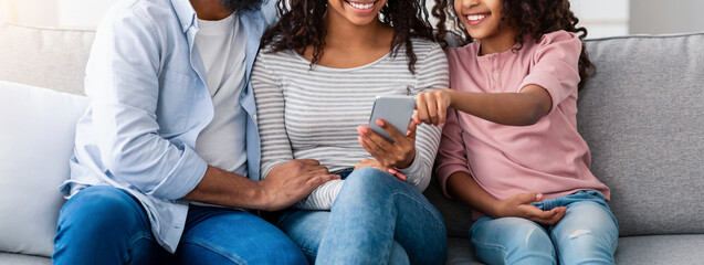 People And Technology. Portrait of happy African American family holding and using mobile phone sitting on the couch at home. People spending time together, sharing social media, watching funny video