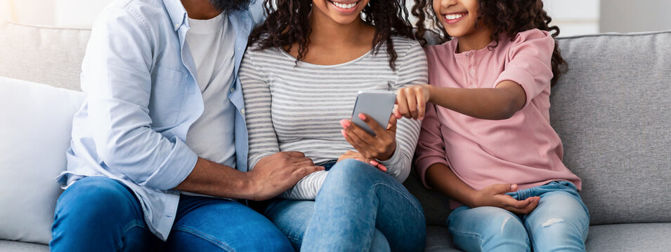 People And Technology. Portrait of happy African American family holding and using mobile phone sitting on the couch at home. People spending time together, sharing social media, watching funny video - Powered by Adobe