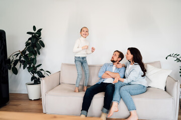 Obraz premium Playful girl stands on couch while parents respond with humor and warmth. Family’s relaxed digital lifestyle shown through casual body language and hidden tech nearby.