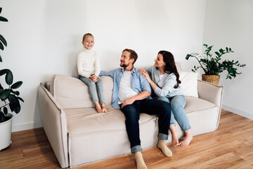 Mother reaches for a mug on a kitchen shelf while father and daughter study together at a wooden table with a tablet. Emphasizes merged roles: educator, nurturing parent, digital learning.