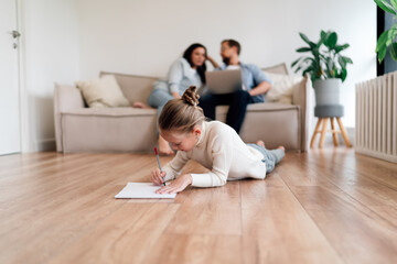 Girl draws on floor with parents in background using laptop, illustrating balance between analog creativity and digital engagement in tech-aware family home.