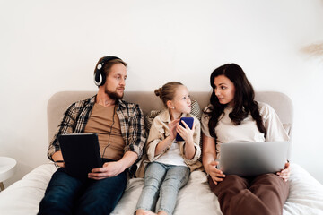 Family sits together in bed using individual gadgets, daughter between parents, symbolizing tech inclusion, curiosity, and shared digital environment across generations.