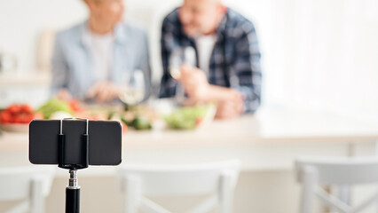 Modern food blog and video review about food delivery service. Smartphone on tripod shoots happy middle-aged man and woman with wine, talk in modern kitchen interior with table with fresh vegetables