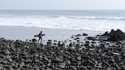 A surfer crosses a boulder strewn beach to get to the waves.