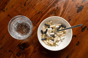 Assorted dietary supplements in a bowl with a spoon beside a glass of sparkling water on a wooden table, promoting health and wellness lifestyle
