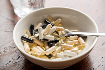 Assorted dietary supplements in a bowl with a spoon beside a glass of sparkling water on a wooden table, promoting health and wellness lifestyle
