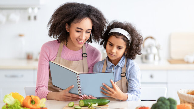 Time With Family. Portrait of African American little girl and young woman holding cookbook, reading and looking for new recipe, cooking dinner in the kitchen, mum and daughter wearing aprons