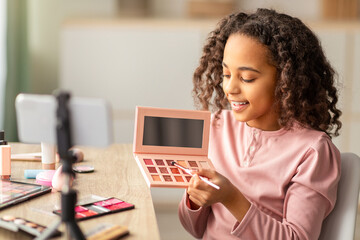 African american young beauty blogger doing makeup and recording vlog, showing her palette to smartphone camera. Little female vlogger talking in front of phone on tripod at home