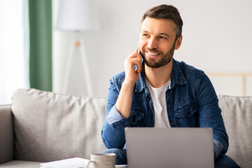 Smiling businessman working with laptop and having conversation on mobile phone, home interior, looking at copy space. Happy middle-aged man working from home, talking to clients on smartphone