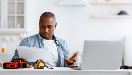 Problems with instructions. Puzzled black handyman reading manual and does not understand what to do, sitting at table with tools and laptop in kitchen interior. Home repairs concept