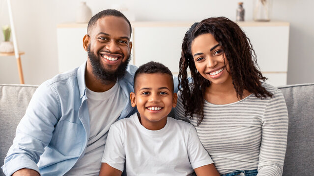 Happy Loving Family. Portrait of cheerful African American son sitting on the couch between his smiling parents. Positive woman, man and boy posing for photo and looking at camera at home
