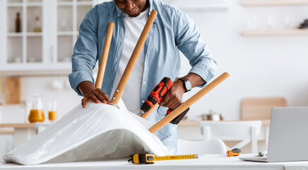 Repair or furniture assembly process. Black craftsman screwing legs to chair with electric screwdriver in kitchen interior, panorama. Unrecognizable man fixing armchair at home