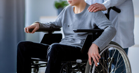 Cropped view of nurse supporting handicapped teenage boy in wheelchair at home, closeup. Unrecognizable impaired adolescent suffering from depression, receiving medical help © Prostock-studio