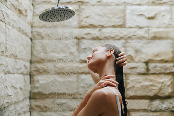 Relaxed woman taking outdoor shower with stone wall background, water running, natural light, casual, calm mood, and wet hair.