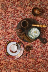 Coffee table setting with white cups, red and gold bordered plates on oriental patterned tablecloth.