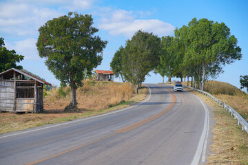 a rural or semi-rural scene featuring a winding road ascending a gentle hill. On the left side of the road, there is a small, weathered wooden structure, possibly a shack or a small dwelling. Trees ar