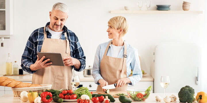 Homemade food and prepare holiday dinner for family at home together. Smiling senior man looks in tablet, wife prepares salad in modern kitchen interior with table with fresh vegetables, copy space