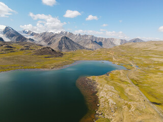 A breathtaking aerial view of a tranquil alpine lake surrounded by green tundra, with a stunning backdrop of rugged, snow-capped mountain peaks