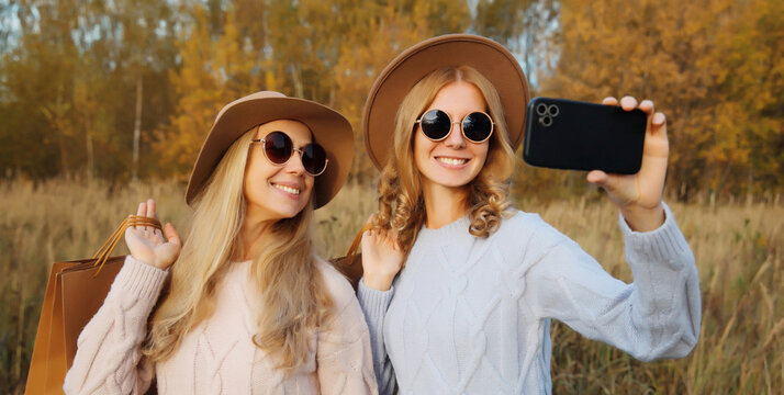 Happy women friends taking selfie with phone, shopping bags in autumn, girlfriends smiling in park - Powered by Adobe
