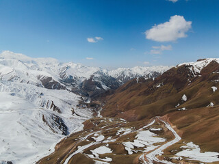 A dramatic aerial view of a mountain valley where snowy peaks transition to brown, earthy slopes, with a winding road and a village visible below