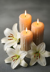 Three burning candles are surrounded by white lilies resting on a textured gray surface
