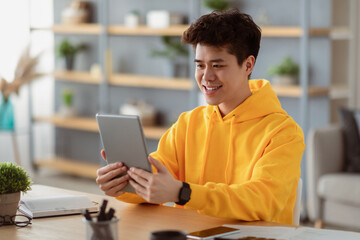 Fototapeta premium People And Technology. Portrait Of Smiling Asian Man Holding And Using Digital Tablet Sitting At Desk Indoors In Living Room. Happy Male Making Video Conference, Studying Or Working Remotely
