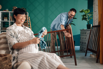 Father assembling baby crib while pregnant wife holds baby clothes