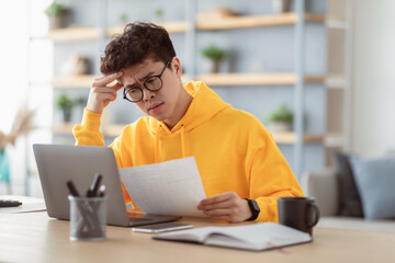 Fototapeta na wymiar Paperwork Concept. Portrait of focused asian man in eyeglasses holding reading paper or financial document, sitting at desk with laptop computer in living room, thinking and working at home office