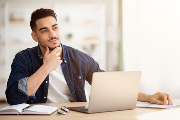 Fototapeta premium Dreamy young arab man sitting at office, looking at copy space, working on laptop, taking notes. Handsome middle-eastern guy dreaming about prosperous future and successful career