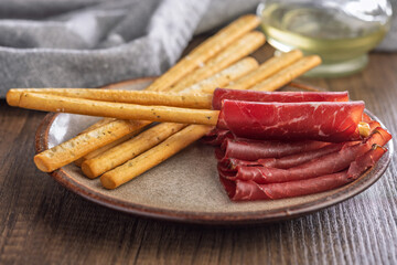 Slices of smoked bresaola and grissini sticks on plate on wooden table.